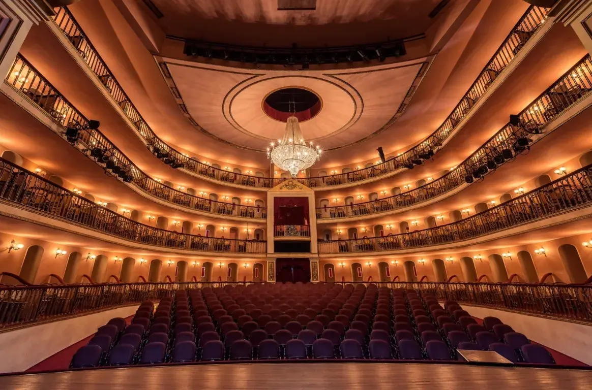 A sala do Teatro São Luiz vista do palco, com plateia de cadeiras roxas, várias ordens de balcões iluminados e um lustre de cristal ao centro.